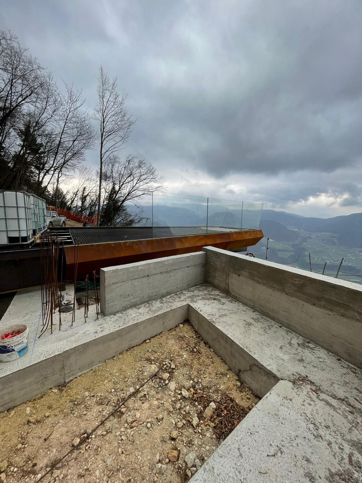 Una terrazza sopraelevata in costruzione con pareti in cemento e ringhiera in vetro che si affaccia su una catena montuosa.