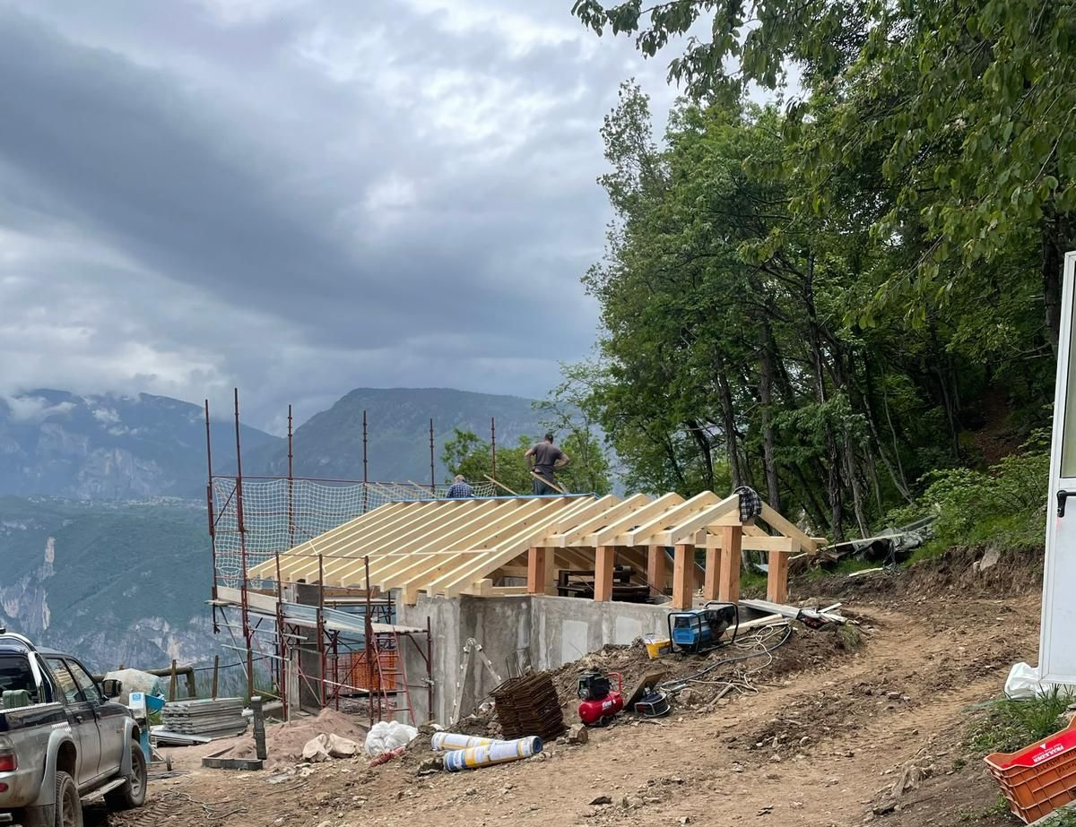 Un operaio si trova in piedi sulla struttura in legno di una casa in costruzione su un pendio che domina le montagne.