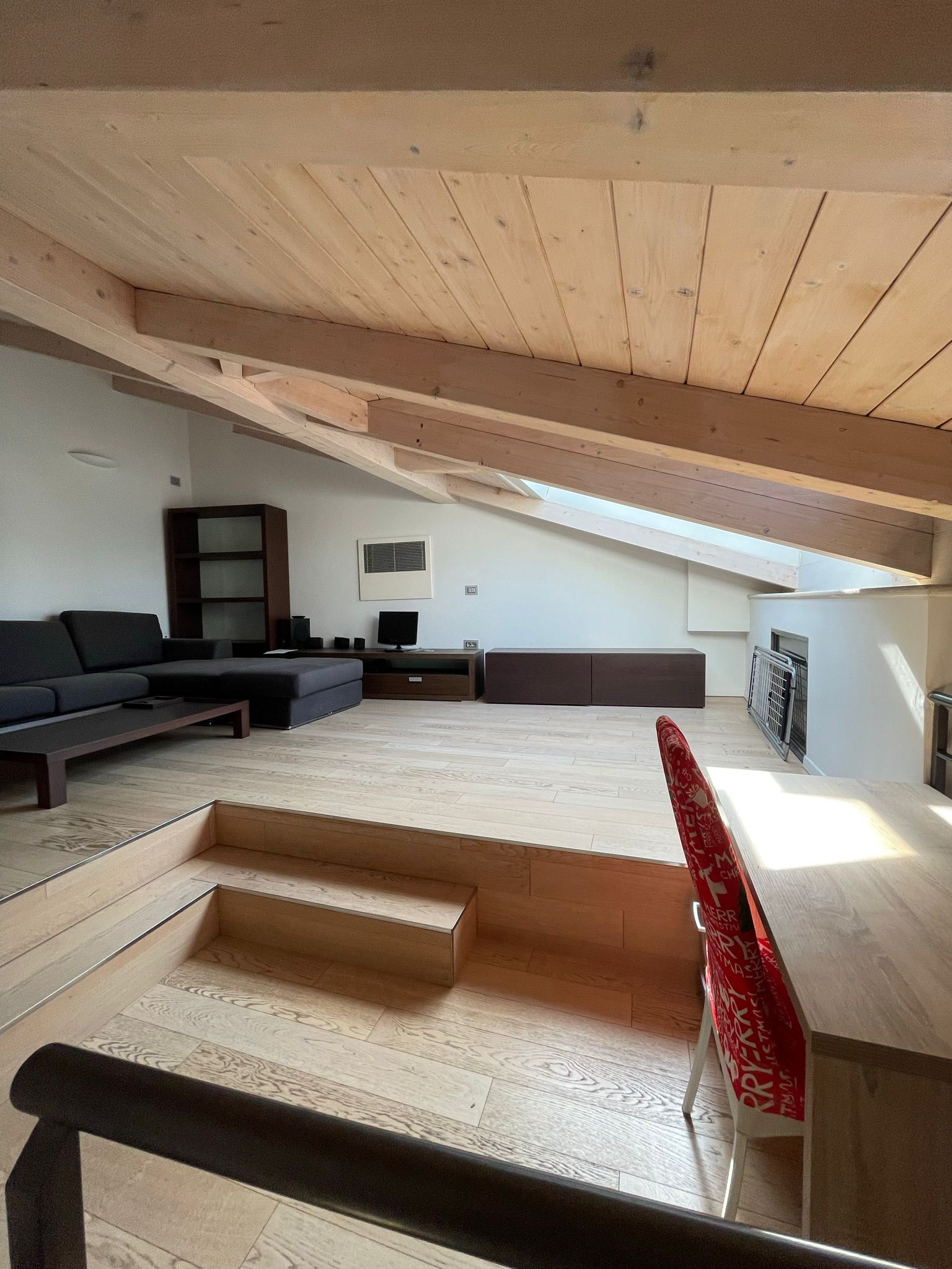 Una mansarda open space con soffitto in legno inclinato, zona divano e scrivania con sedia rossa a fantasia su un ripiano rialzato.