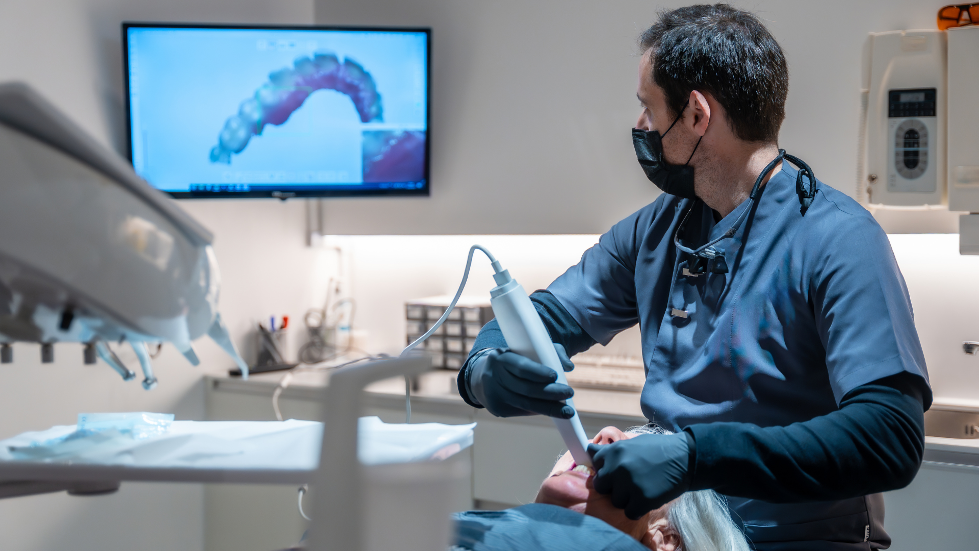 Dentist wearing a mask using a scanning tool on a patient; a digital image of teeth is displayed on a monitor.