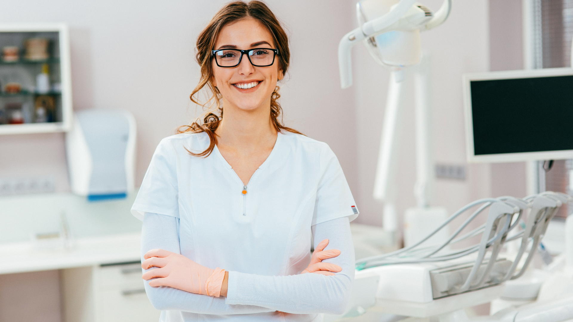 A smiling dental professional in a white uniform stands with arms crossed in a clinical office setting.