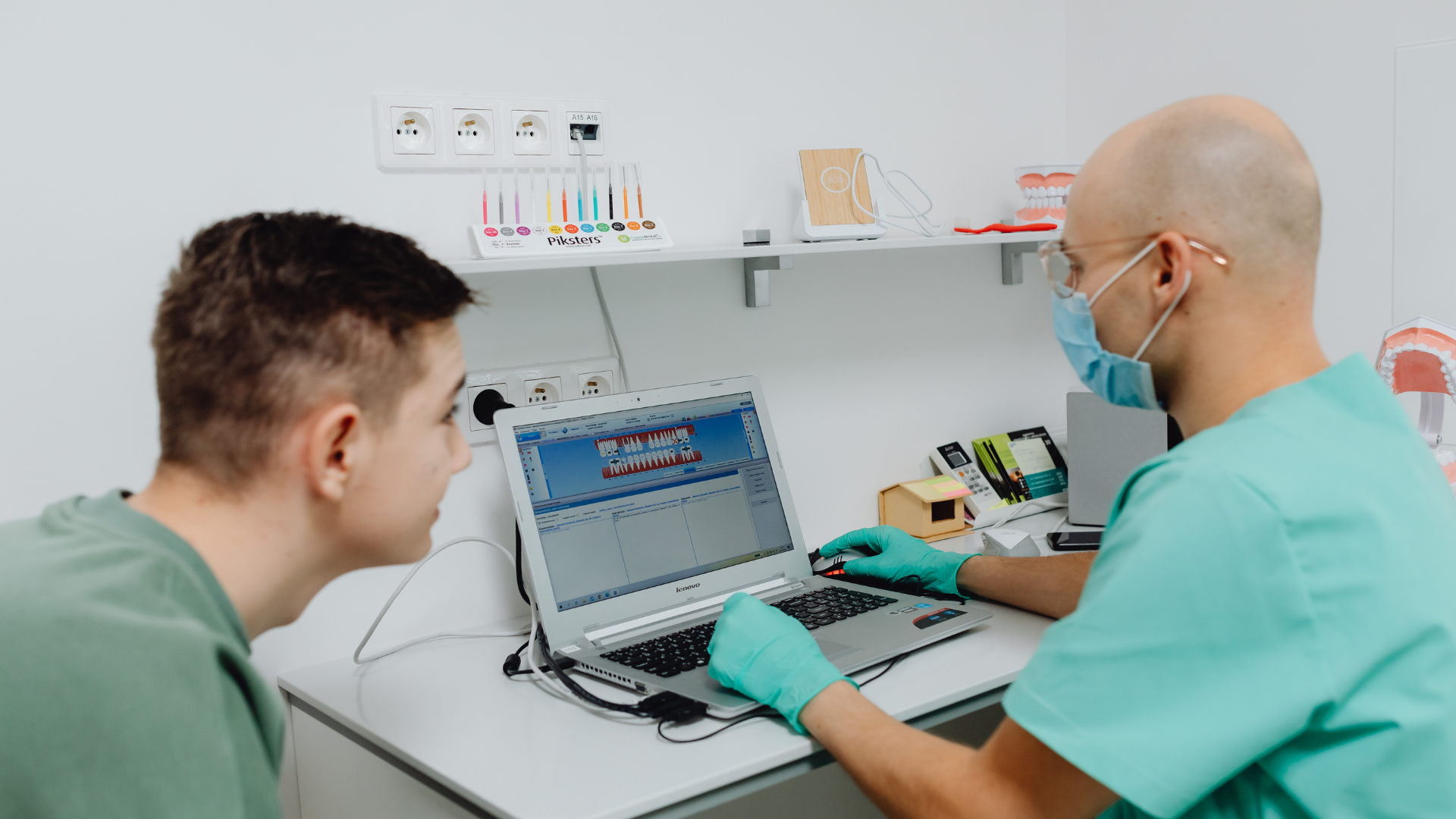 Dentist in surgical scrubs, mask, and gloves showing a patient an x-ray on a laptop.