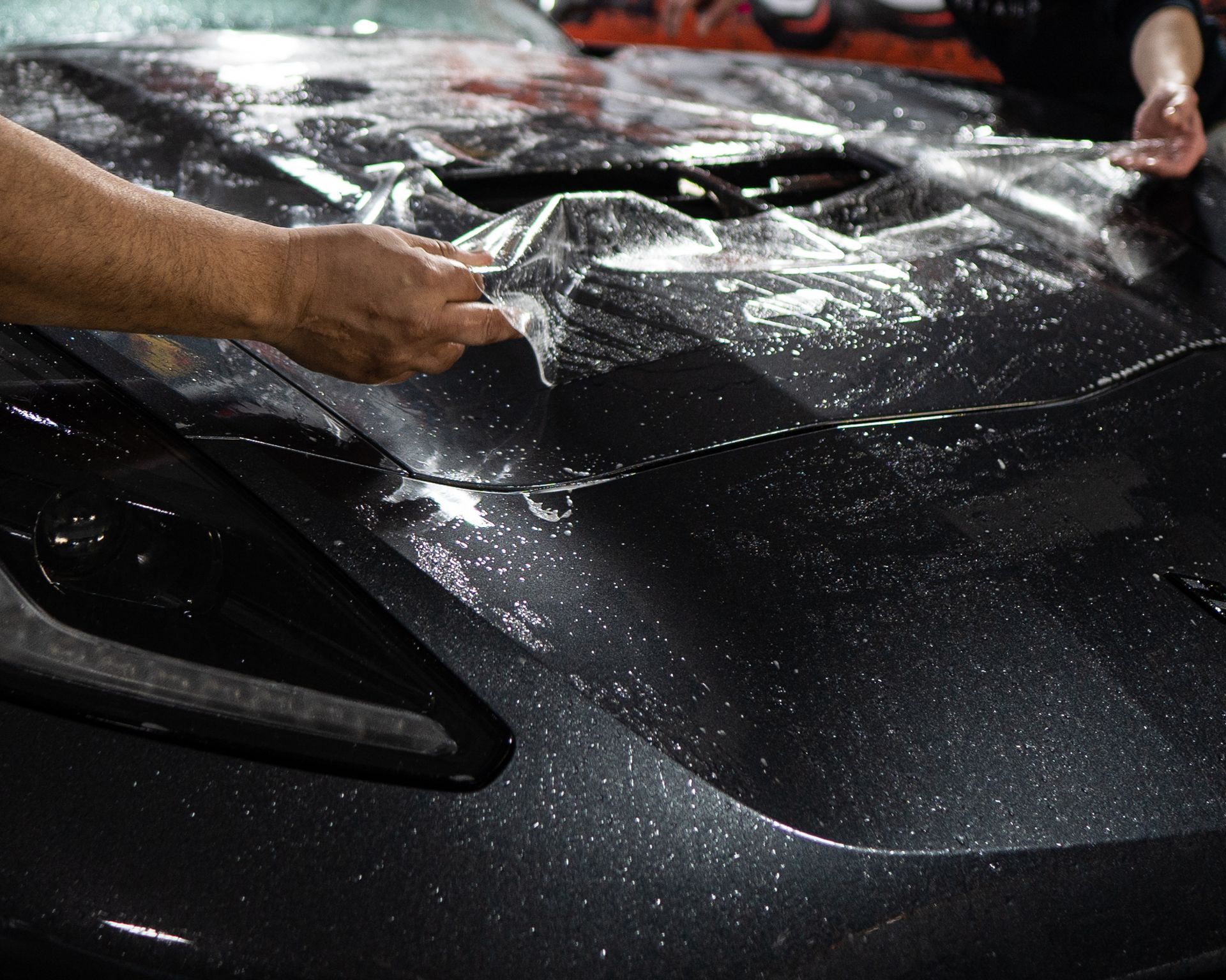 A person is covering the hood of a black car with plastic wrap.