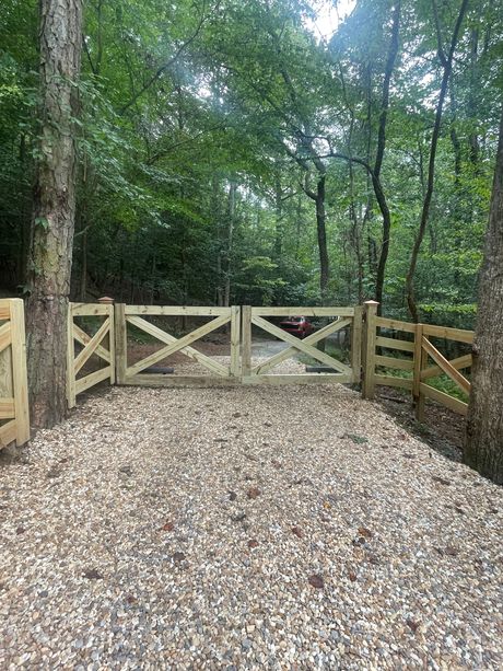 A split rail wooden fence surrounds a gravel driveway in the woods.