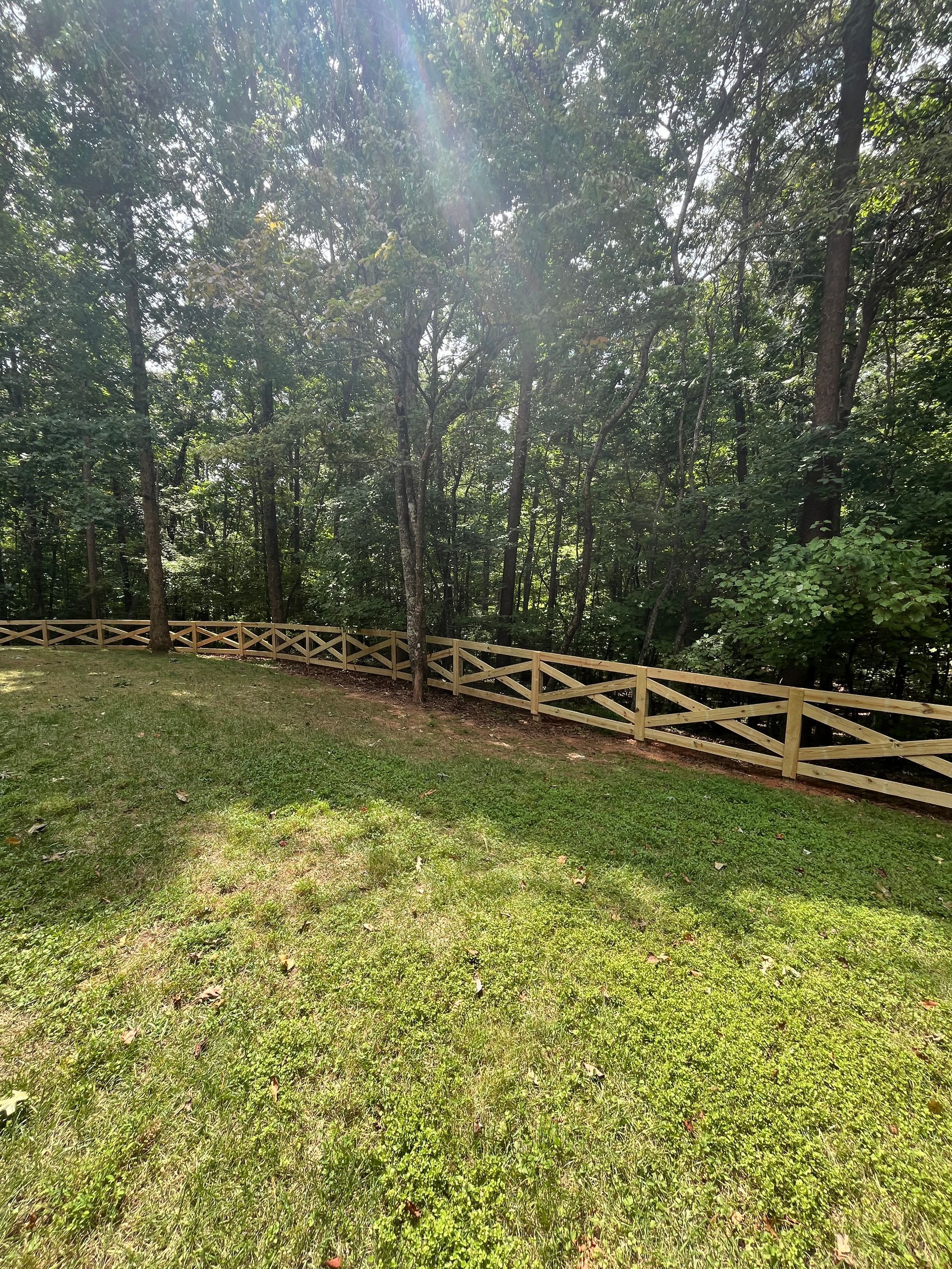 A splt rail wooden fence surrounds a lush green field with trees in the background.