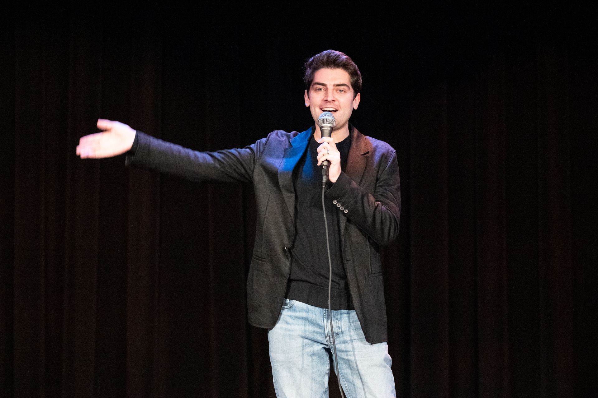 A man comedian is standing on a stage holding a microphone.