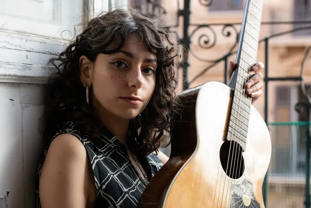 Woman with curly hair holds a guitar, leaning against a window, looking at the camera.