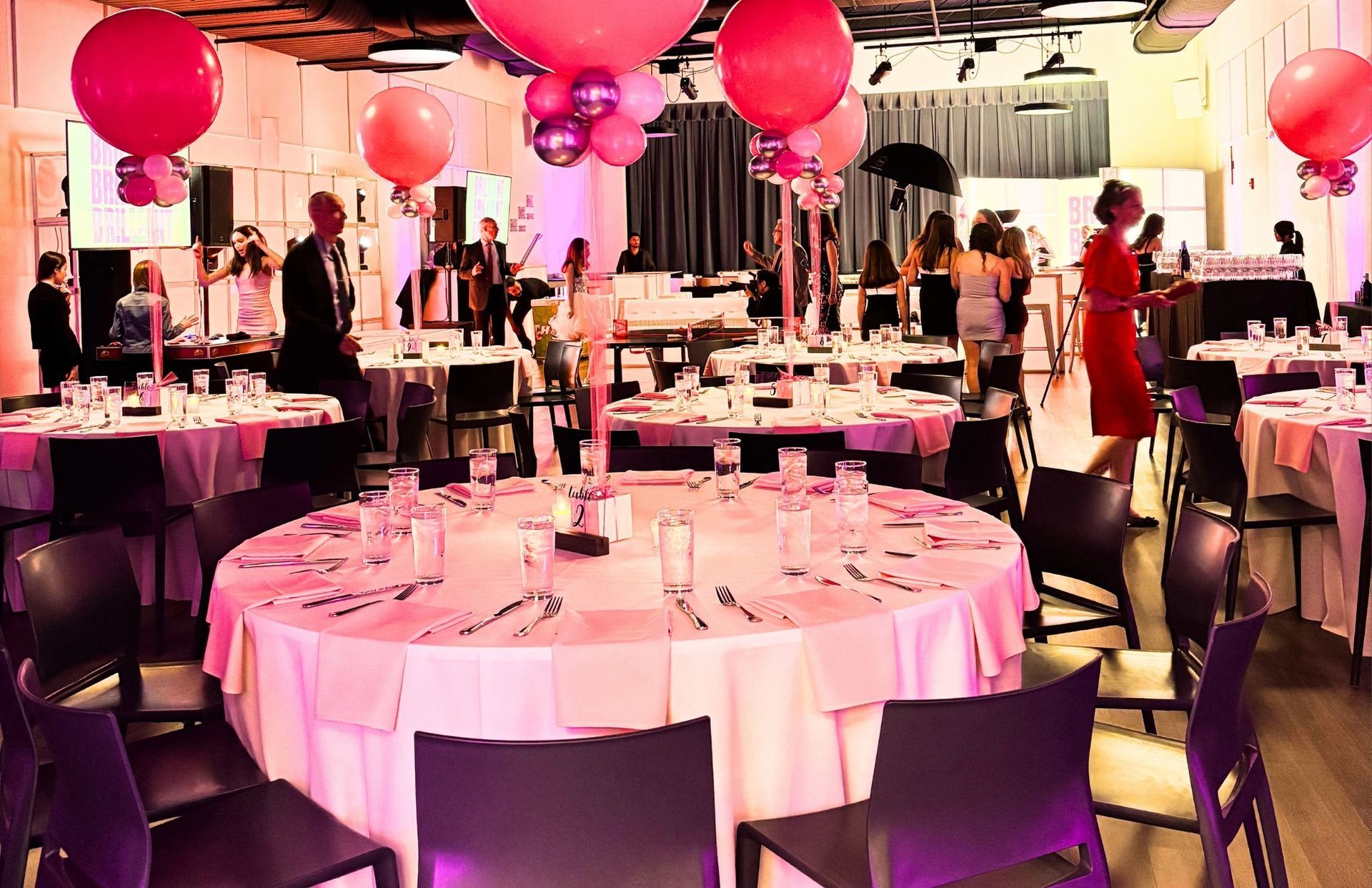A large room with tables and chairs set up for a party with pink balloons hanging from the ceiling.