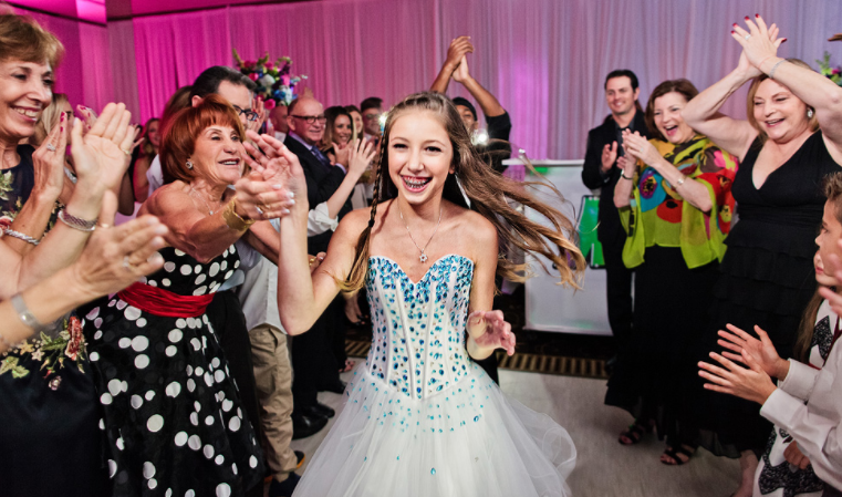Girl in blue and white dress, surrounded by clapping people at a party, celebrating.