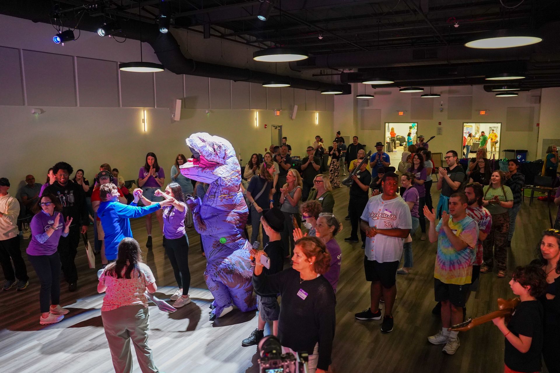 A group of people are dancing in a large room with a dinosaur costume.
