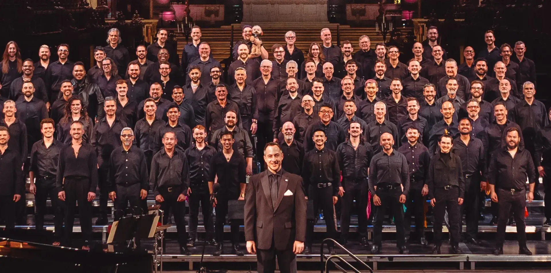 A large choir in black attire, led by a conductor in a suit, on a stage with a dark backdrop.