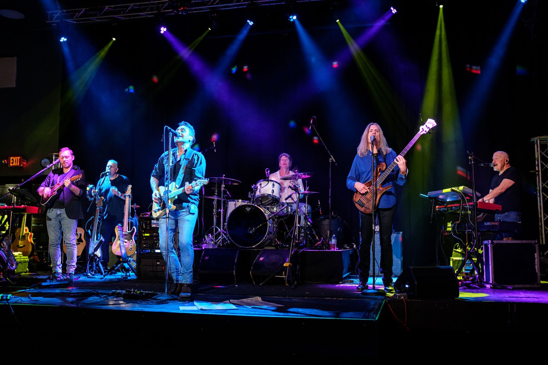 Band performing on stage, lit with blue, green, and purple lights. Musicians play instruments, singing into microphones.