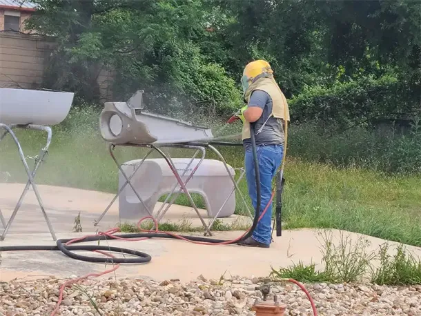 A worker in a white hazmat suit and yellow hard hat uses a power tool to cut into the brick wall of a beige building.