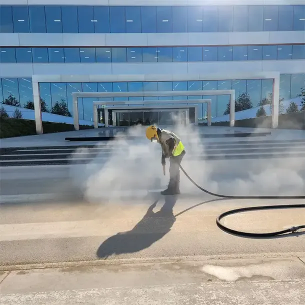 A worker in a yellow safety vest uses a power sprayer to clean the concrete ground in front of a modern glass building.