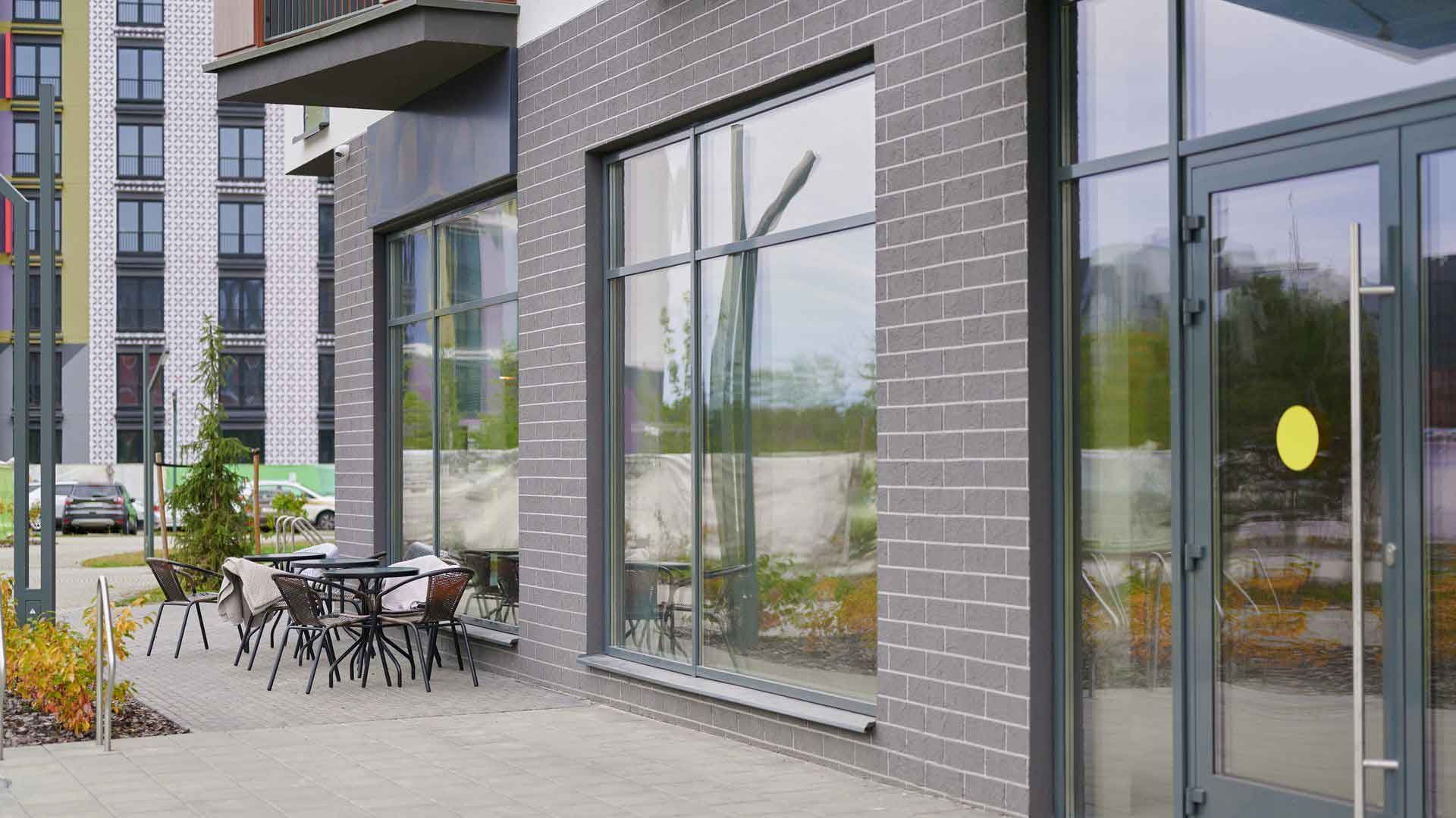 Exterior of a building with large windows and a seating area outside. Gray brick facade, glass doors, and tables/chairs.