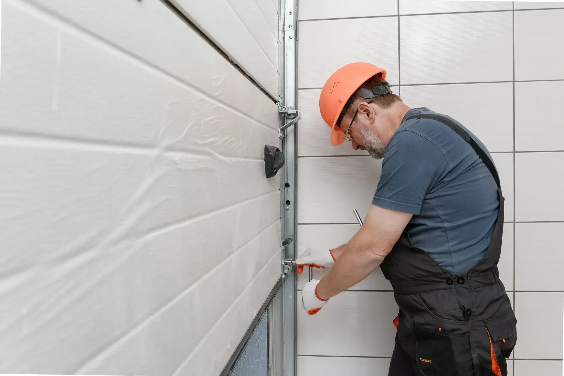 A person in overalls and a hard hat is repairing a white garage door with tools.
