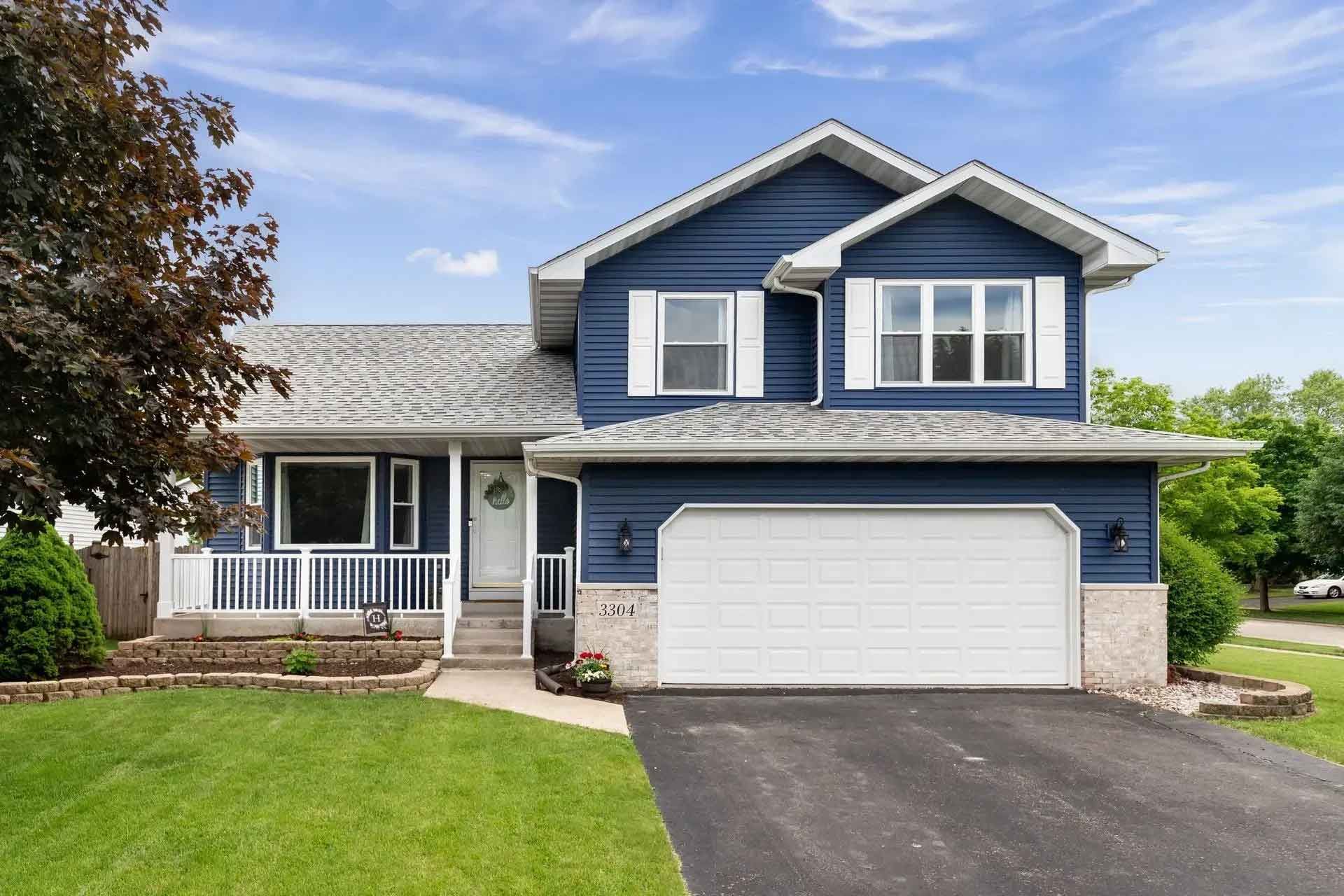 Blue two-story house with white trim, a two-car garage, and a green lawn under a blue sky.