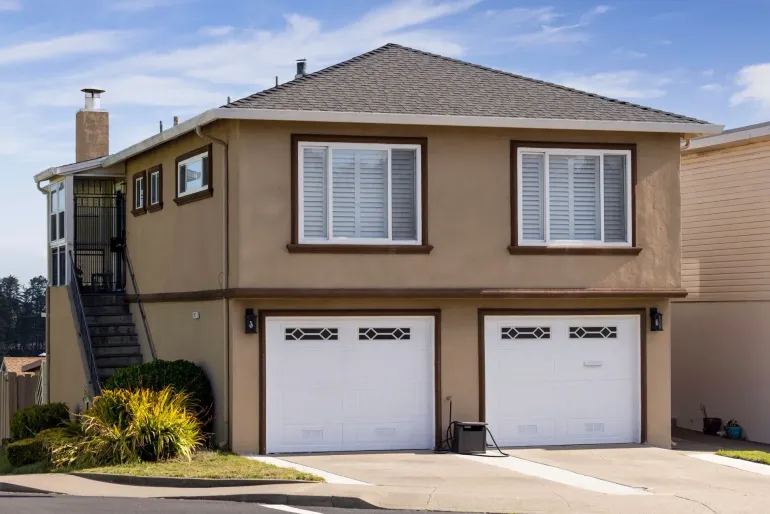 Two-story beige house with white garage doors and shutters, brown trim, and a staircase on the side.