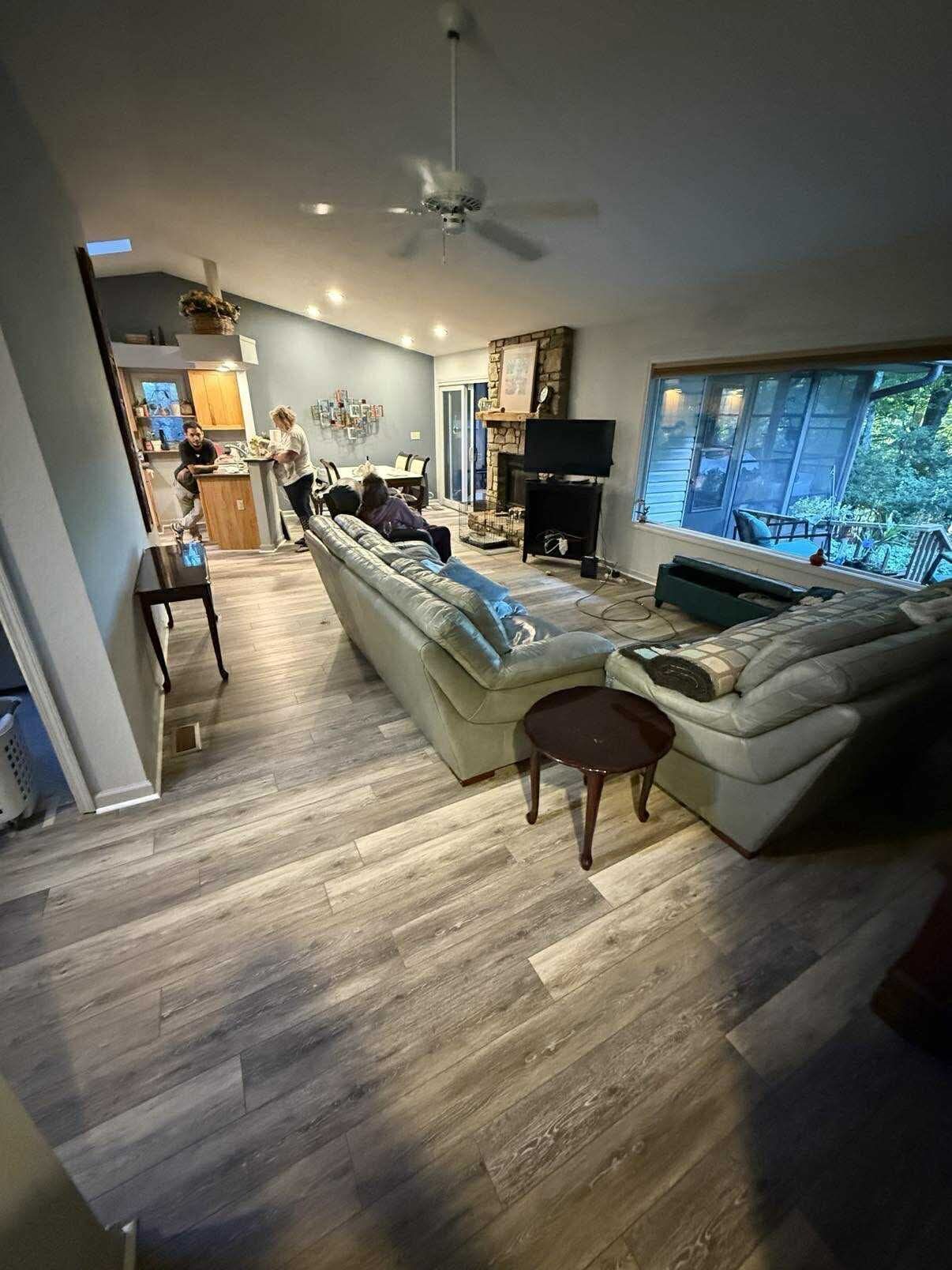 Spacious living room with sectional sofa, fireplace, and large window. Wooden floors. People are present.