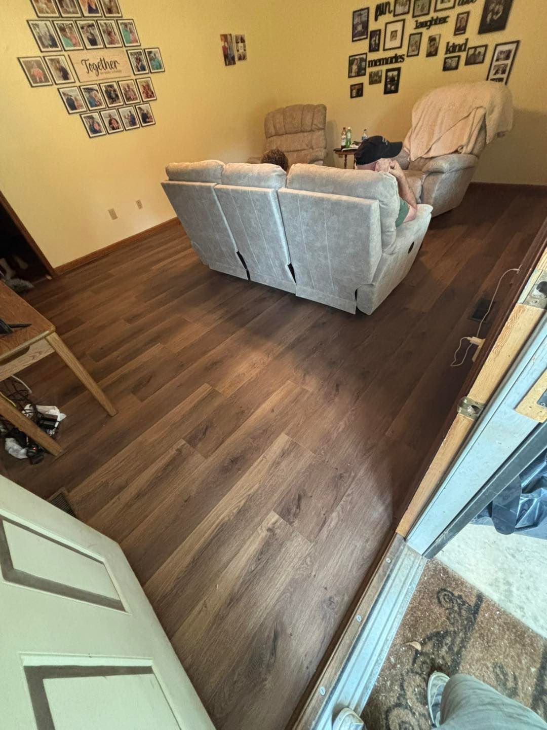 Living room with wood-look flooring, light gray couch, recliner, and photos on the wall.