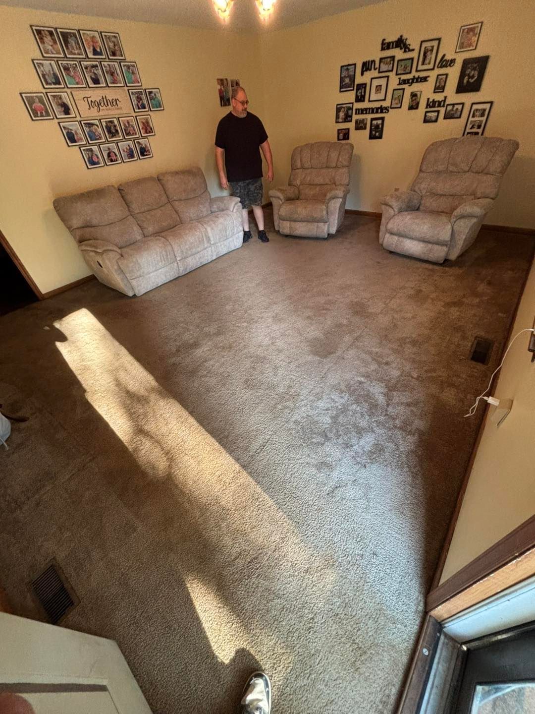 Man stands in a room with a sofa, two recliners, and framed photos on the wall. Brown carpet.