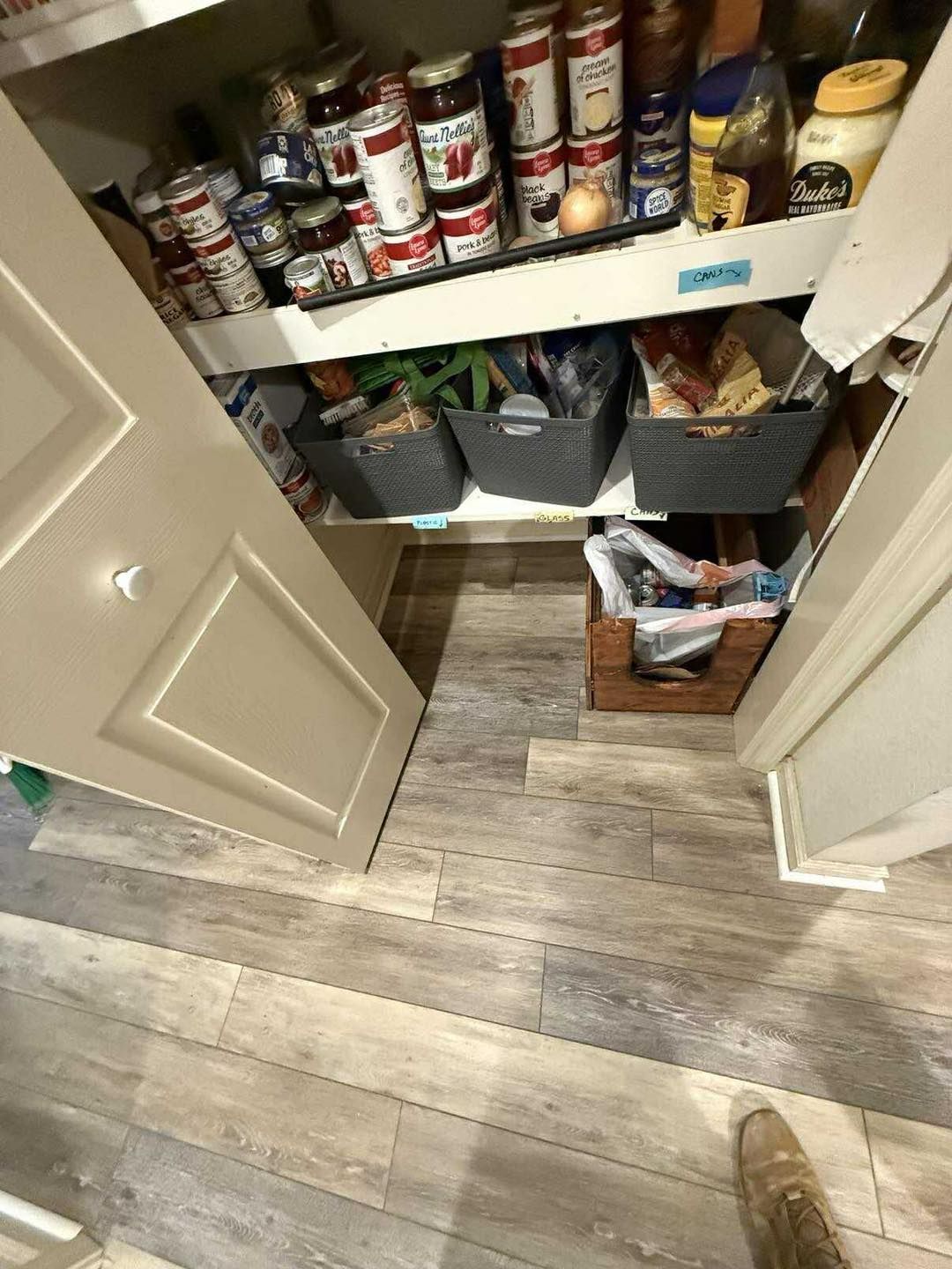 Pantry with shelves of canned goods, storage bins, and a wooden crate; light wood-look flooring.