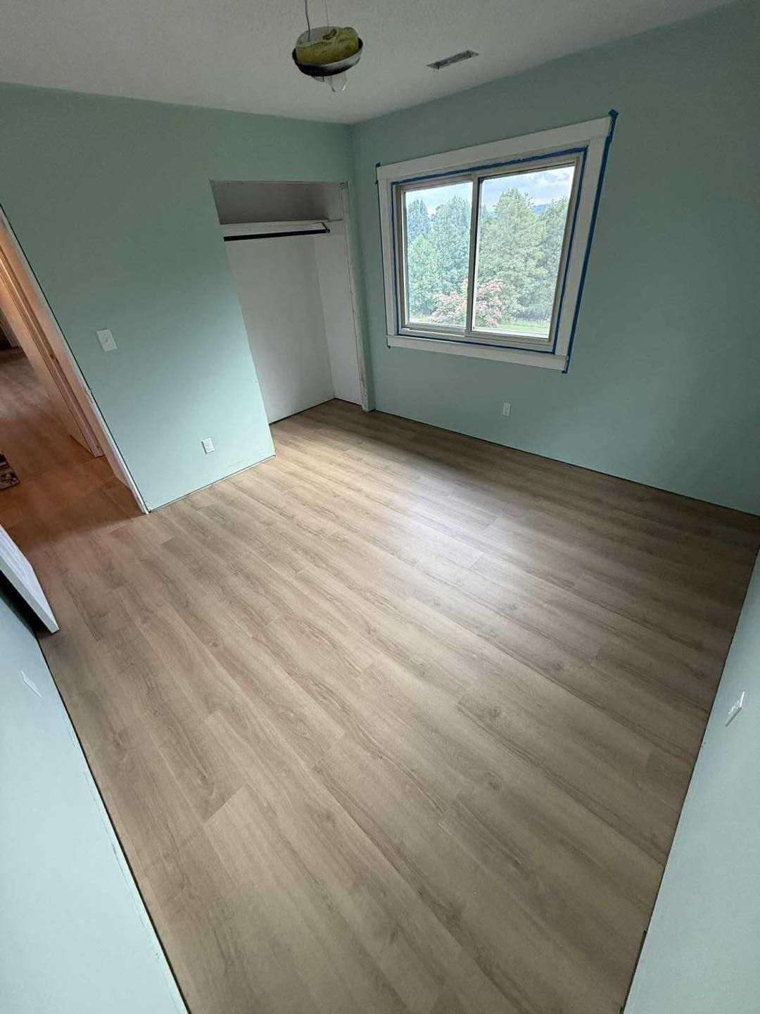 Empty bedroom with light wood-look flooring and light blue walls, window, closet.