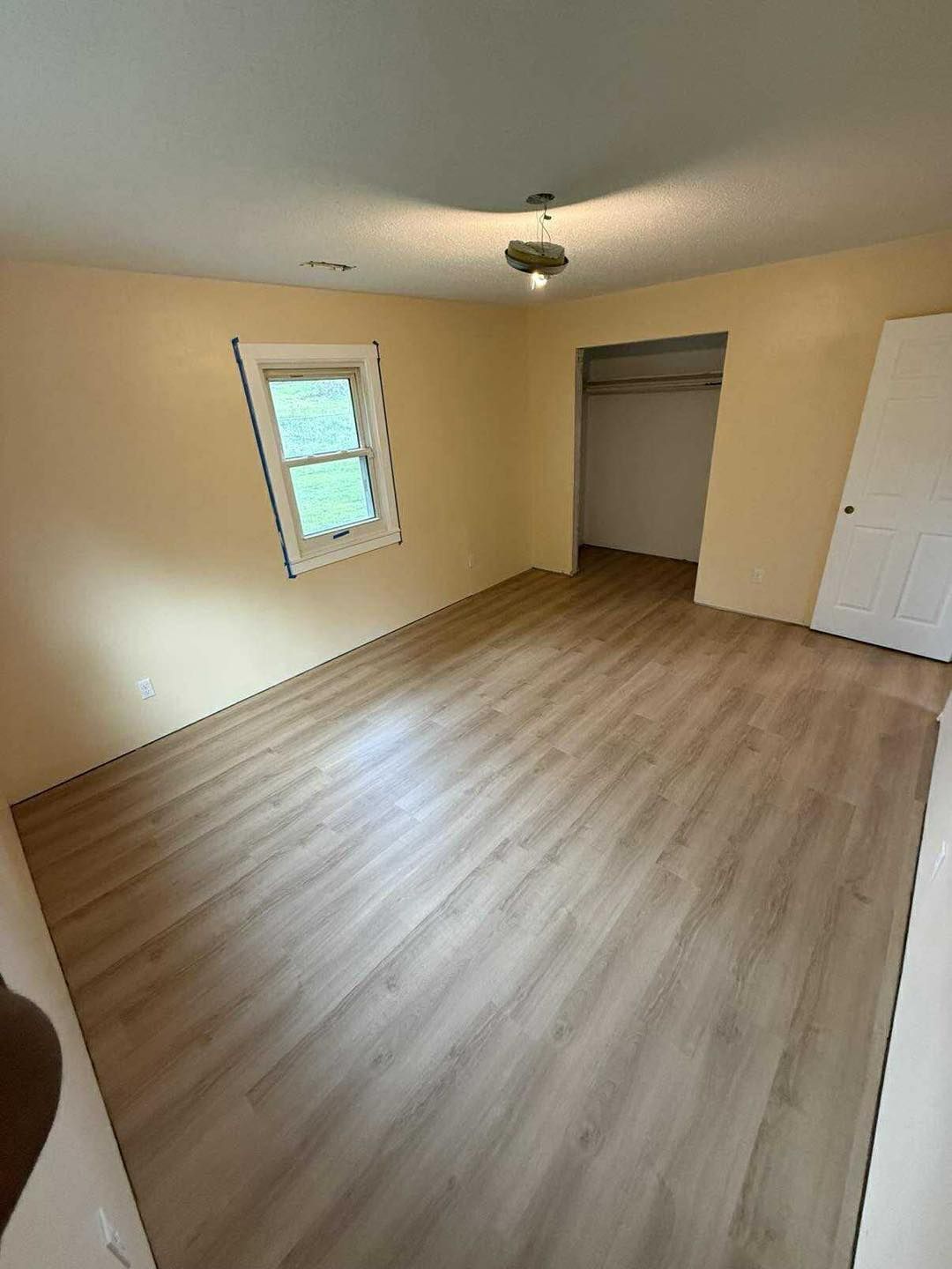 Empty bedroom with wood-look floor, window, closet, and pale yellow walls.
