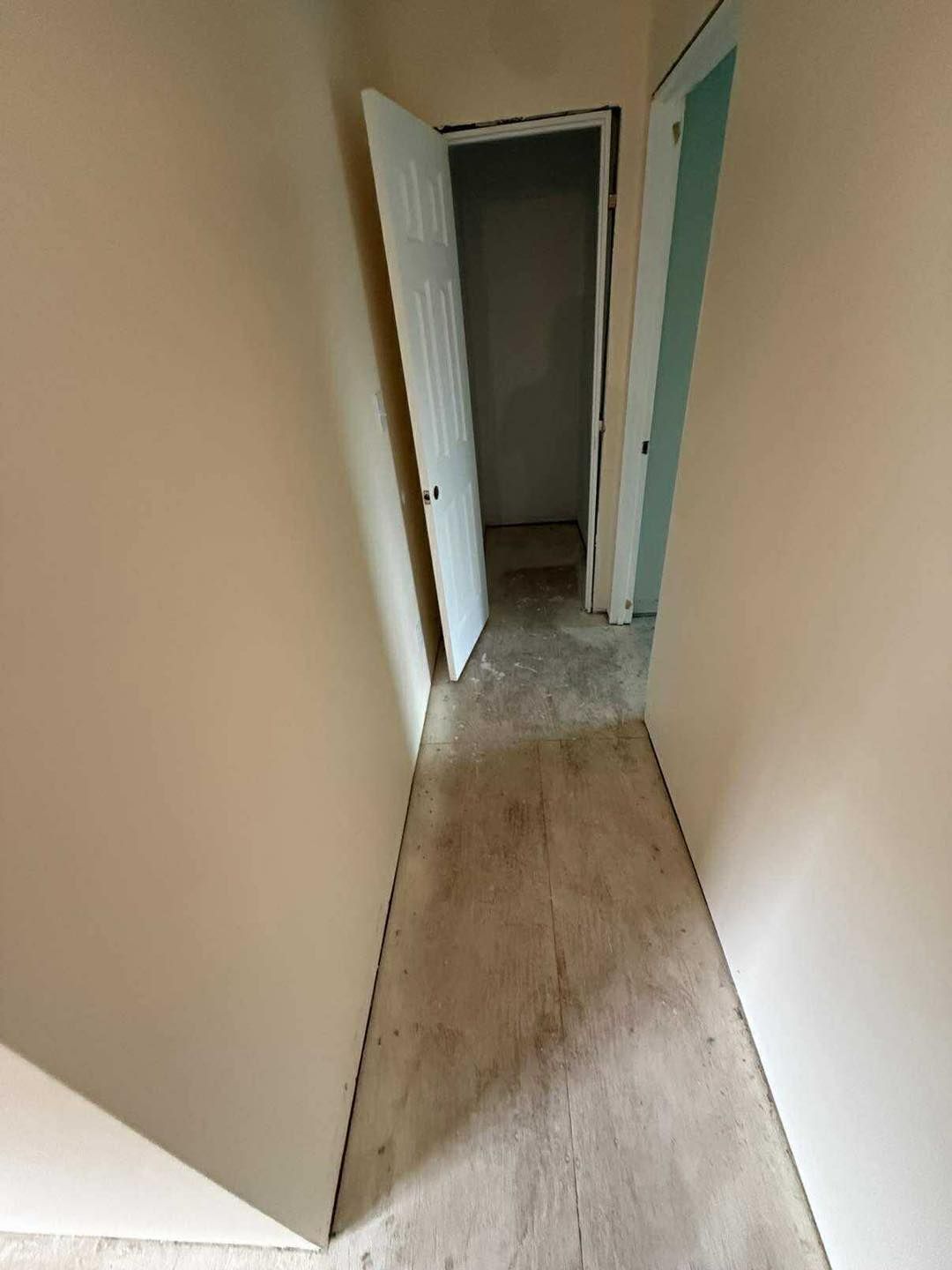 Narrow hallway with beige walls, worn flooring, and a partially open white door leading to a dark room.