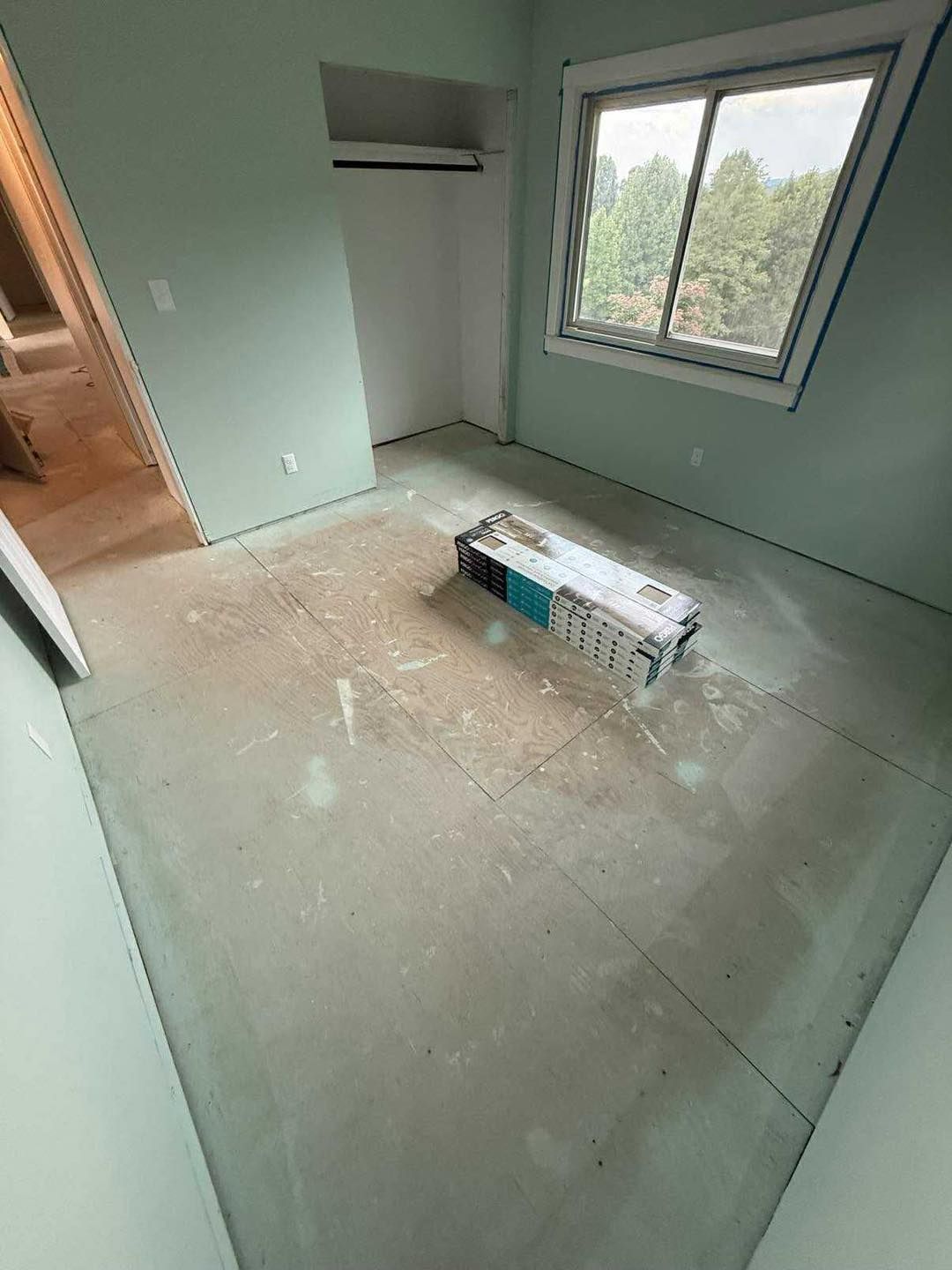 Empty room with light green walls, window, and exposed flooring. A box sits on the floor.