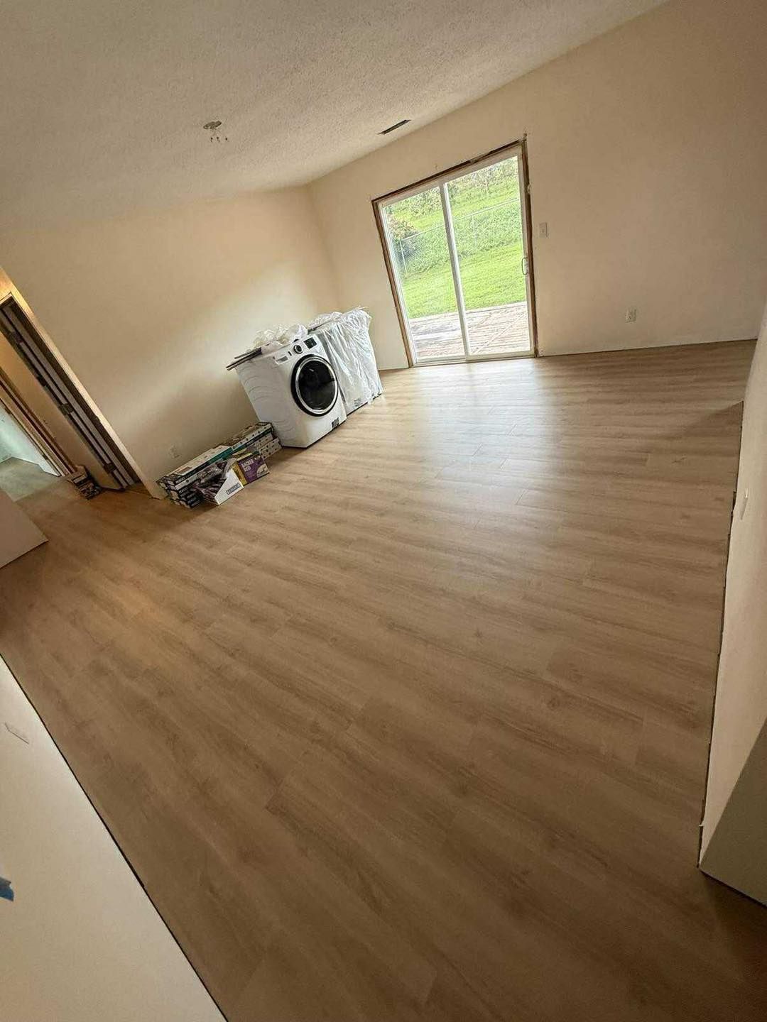 Empty room with new wood-look flooring, beige walls, and a sliding glass door. A washing machine sits inside.