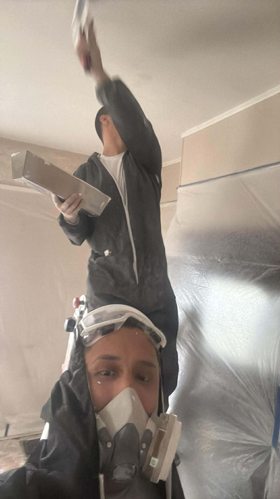 Two people renovating a room. One on a ladder, plastering ceiling. Another wears a respirator, taking a selfie.