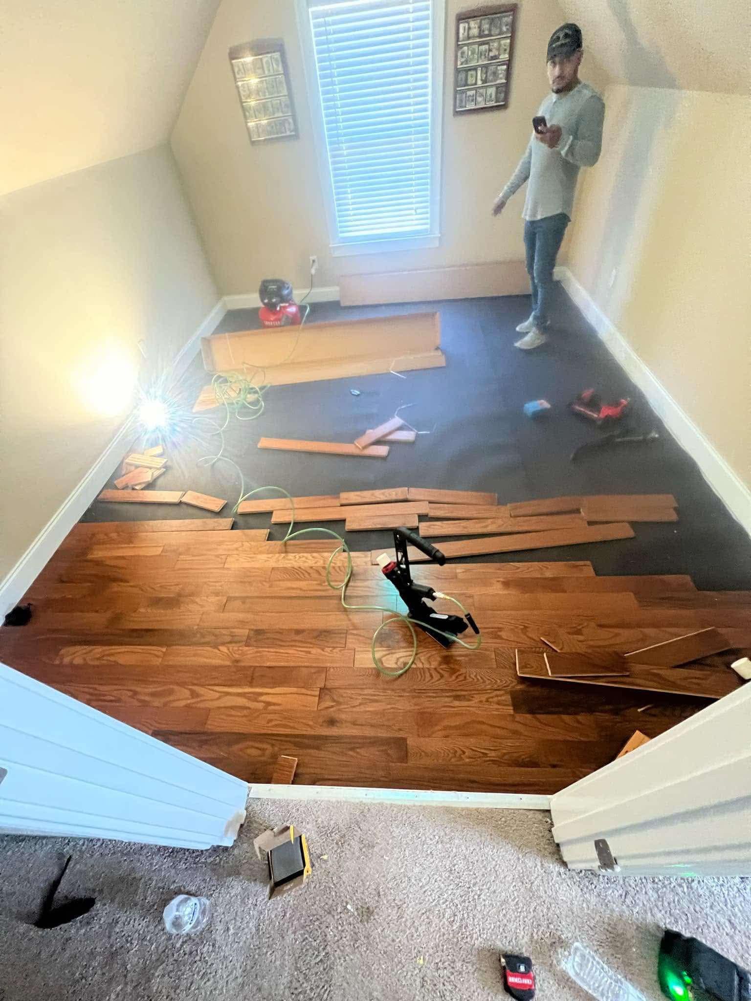 Man installing hardwood flooring in a room with a window, tools, and materials.
