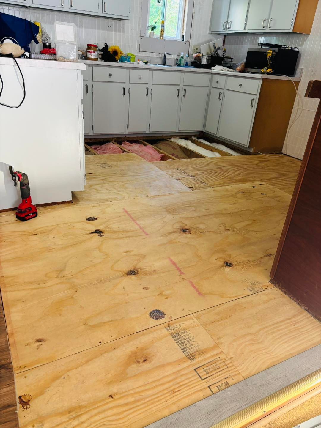 Kitchen with exposed plywood subfloor; cabinets, window, refrigerator in background; construction in progress.