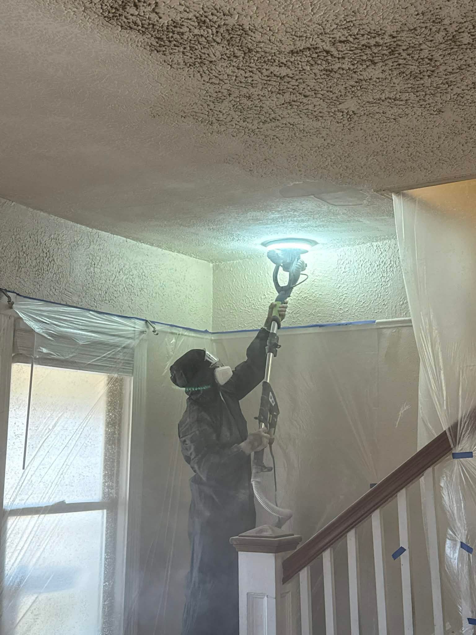 Person in protective gear sanding a moldy ceiling in a room with plastic sheeting and a staircase.
