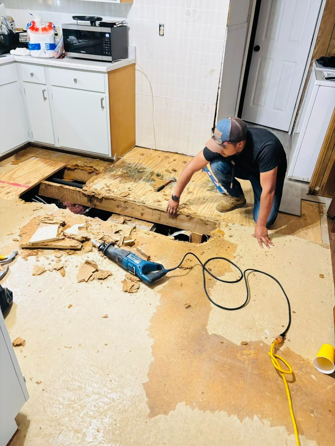 Person examining a hole in a kitchen floor, likely during a renovation. Debris scattered, exposed subfloor.