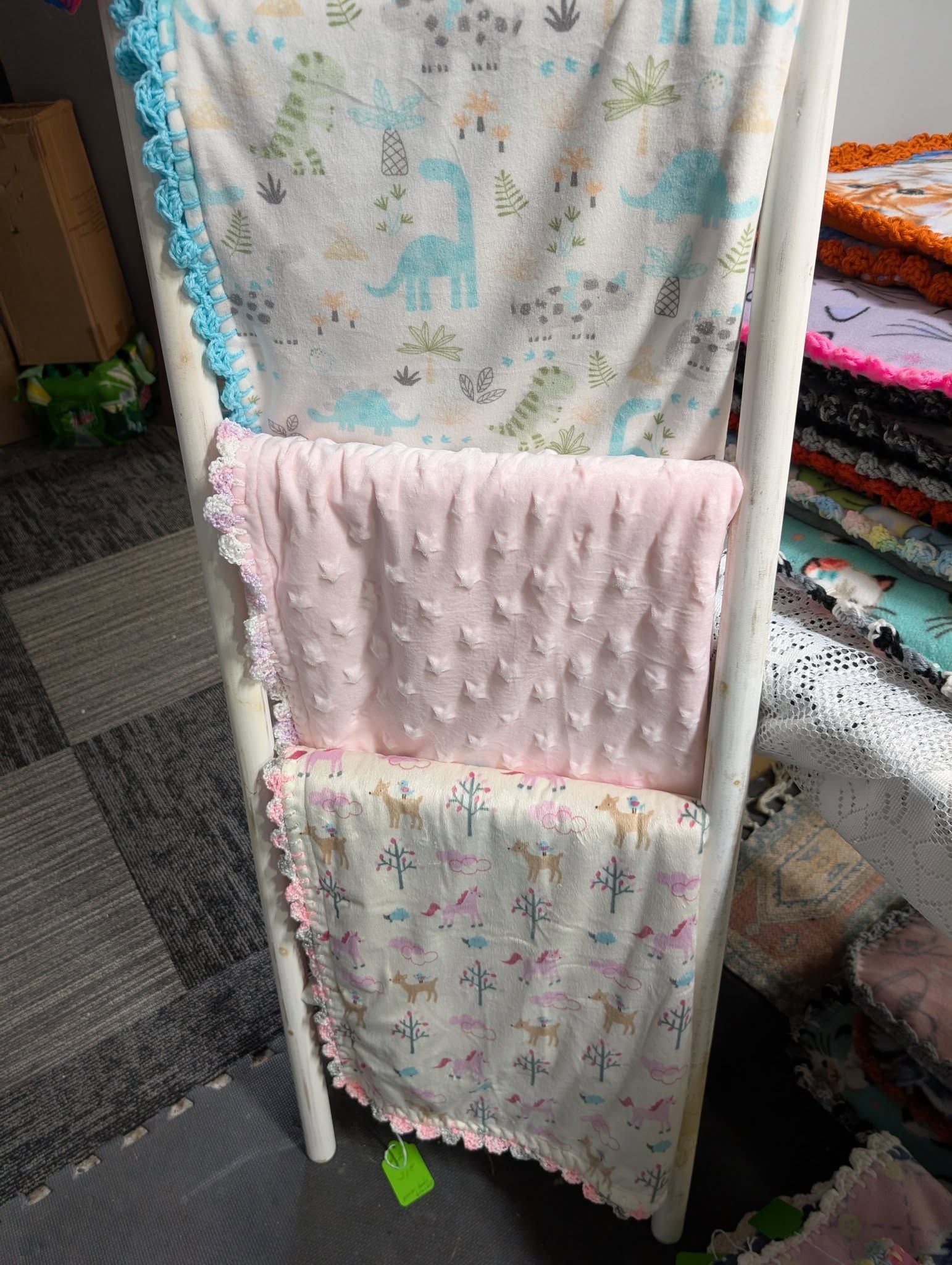 Three baby blankets on a white ladder. Dinosaur, pink textured, and cross print, all with scalloped edges.