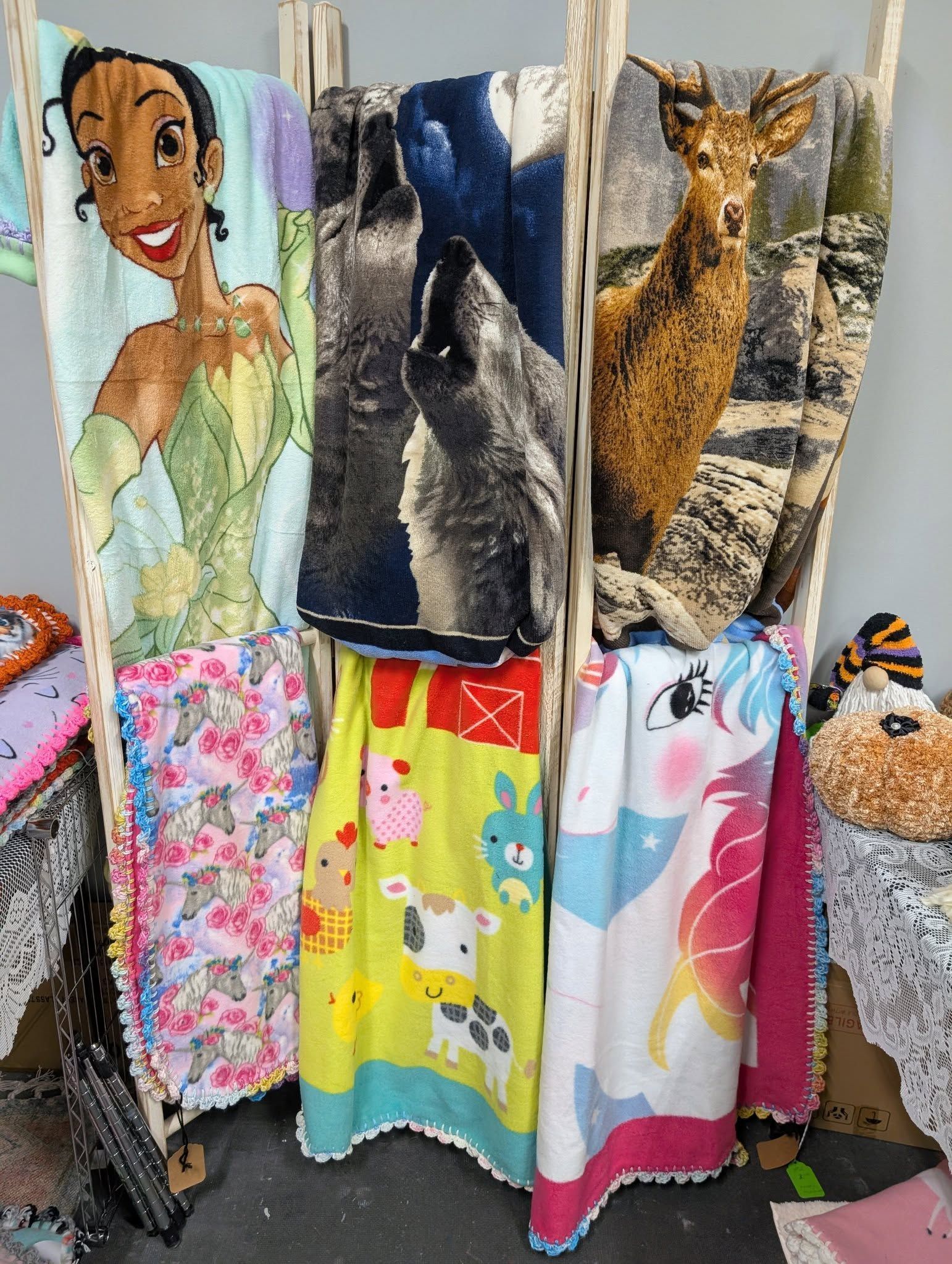 Blankets hanging on a wooden rack; Tiana, wolves, deer, flowers, farm animals, and unicorn designs.
