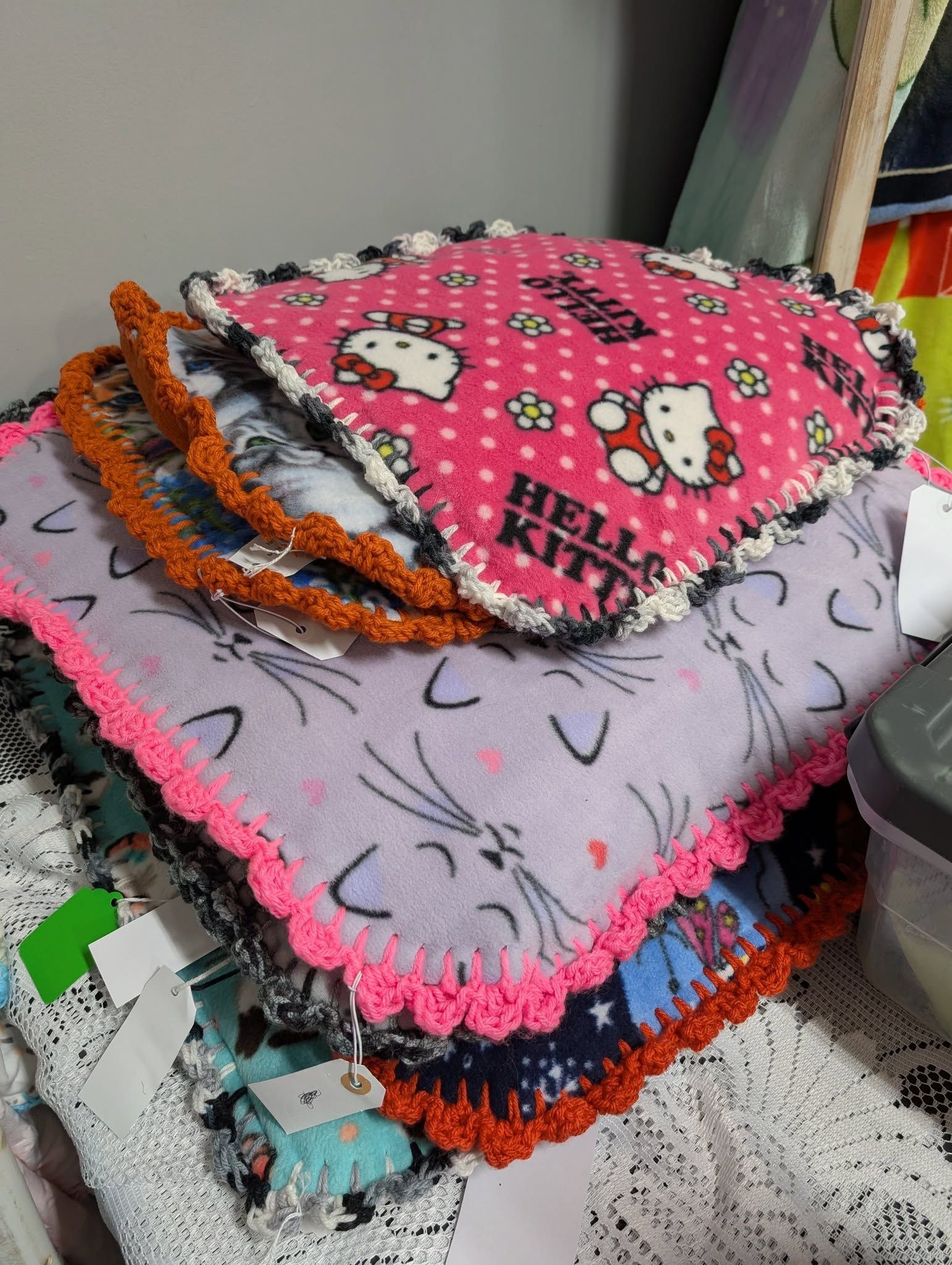 Stack of colorful fleece blankets with crocheted edges, featuring cat and Hello Kitty patterns.