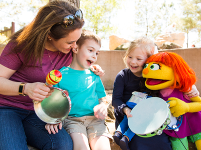 A woman, two children, and a Muppet character sit together outdoors, holding musical instruments and smiling.