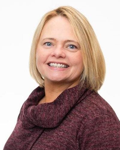 A person with shoulder-length blonde hair, wearing a dark maroon cowl-neck sweater, smiling against a white background.