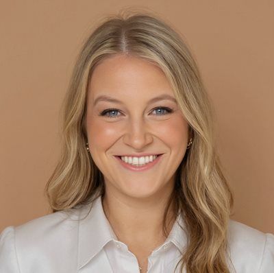 A smiling person with long blonde hair, wearing a white collared shirt, positioned against a plain tan background.
