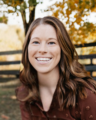A smiling person with long brown hair, wearing a brown shirt, posing outdoors in front of a blurred wooden fence and trees.