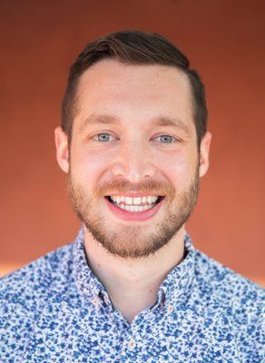 A person with a beard and short hair smiles, wearing a blue and white floral collared shirt against a solid rust background.