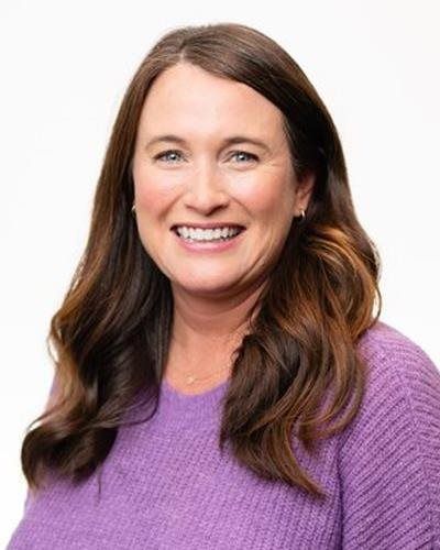 A smiling woman with long brown hair wearing a purple sweater, shown against a plain white background.