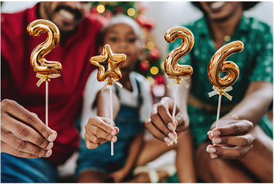 A family holds golden party decorations: a number 2, a star, a 2, and a 6, with a decorated tree in the blurred background.