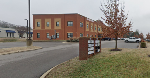 A two-story red brick commercial building with a parking lot and a directory sign on a grassy lawn under an overcast sky.