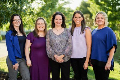 Five women stand side-by-side outdoors, smiling in a park-like setting.