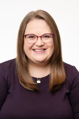 A smiling person with shoulder-length brown hair, wearing glasses and a purple top, against a plain white background.