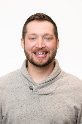A person with short brown hair and a beard, wearing a gray shawl-collar sweater, smiling against a white background.